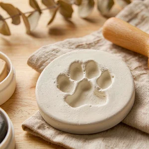 Round clay keepsake disc with a dog paw print impression on linen beside a rolling pin